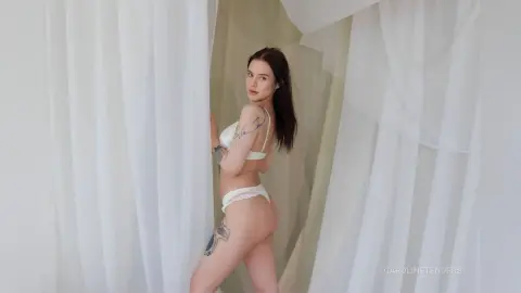 Young woman with tattoos in a white bikini posing between flowing sheer curtains in a bright, airy room.