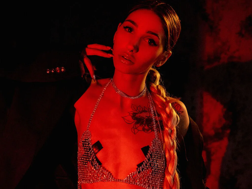 KristiNovak wears a sparkly silver bra and necklace while posing with her blonde braid under red lighting.