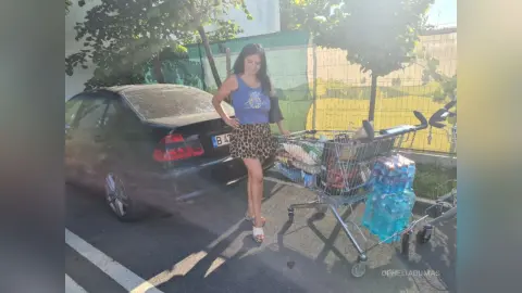 Young woman in a blue tank top and leopard print skirt standing by a car and shopping cart on a sunny day.
