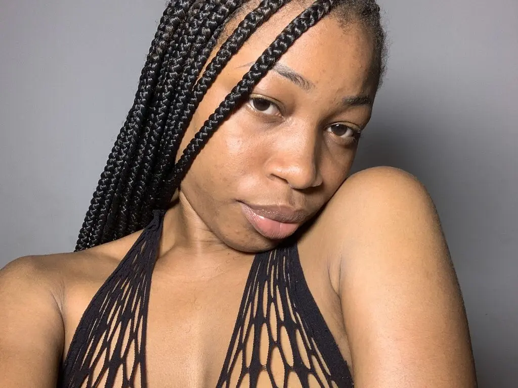 Close-up portrait of a woman with long black box braids wearing a black cutout halter top against a gray wall.