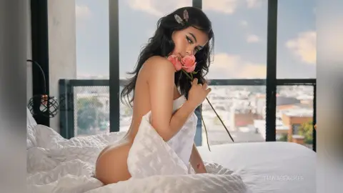 Young woman in white bedsheets holding pink roses on a balcony overlooking a city at sunset.