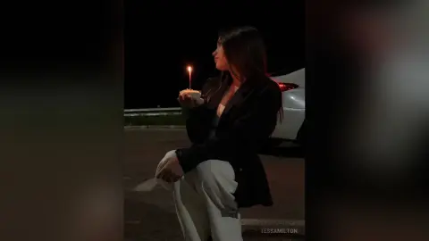 TessaMilton sits outside at night wearing a black blazer and white pants, holding a small cupcake with a single lit candle.