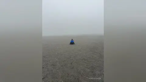 Solitary traveler in a blue jacket sitting on a vast, misty beach under a foggy gray sky.