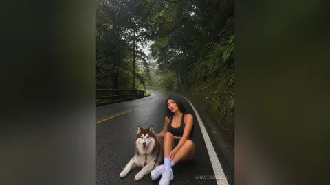 AnastasiaVelo — AnastasiaVelo sits on a paved road next to a large husky dog wearing a black tank top and shorts.