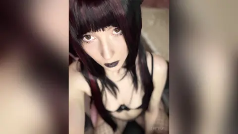 Young woman with dark hair and dark lipstick wearing black lingerie in a soft-lit indoor setting.