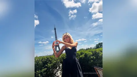 NickyAshcroft poses in a black dress against a blue sky, making a heart shape with her hands in front of the Eiffel Tower.