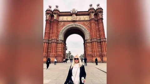 MilenaBia stands in front of a large brick archway wearing a white top and black jacket with sunglasses.