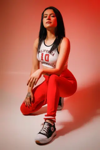 Young woman in white California crop top and red sweatpants posing against a warm red studio background.