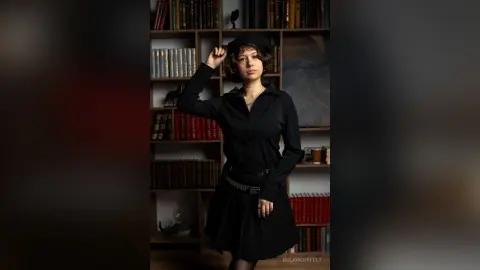 Young woman in a black button-down shirt and skirt posing thoughtfully in a moody, vintage library setting.