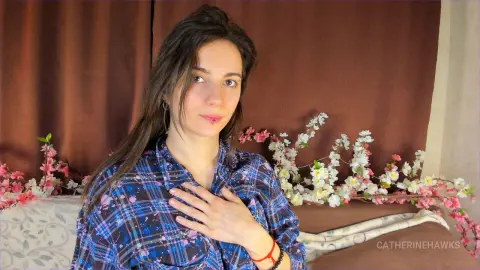 Smiling woman in a blue plaid shirt sitting near pink flowers in a cozy indoor setting.