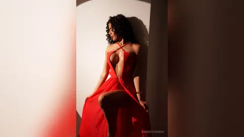 Elegant woman in a flowing red halter dress posing in dramatic sunlight against a white wall.