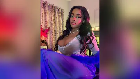 BossyBella — BossyBella poses in a white lace top and a blue skirt, showing off her long wavy hair and pearl necklace in a room with patterned curtains.
