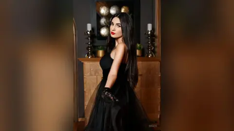 Elegant woman in a black evening gown and lace gloves posing in a moody, luxury interior setting.