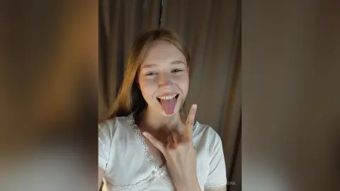 EvelinaMoorina is a blonde woman in a white lace top smiling and sticking her tongue out while making a hand sign.