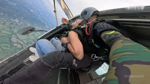 RossiNoleen reclines in the open door of a plane wearing blue jeans and a black harness while skydiving over a green landscape.