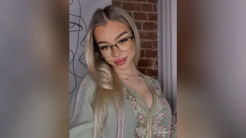 Blonde woman in an embroidered sage green tunic smiling in a cozy room with brick walls.