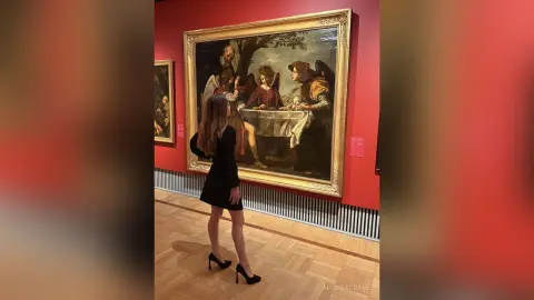 Woman in a black dress admiring a classical Renaissance painting in a red art museum gallery.