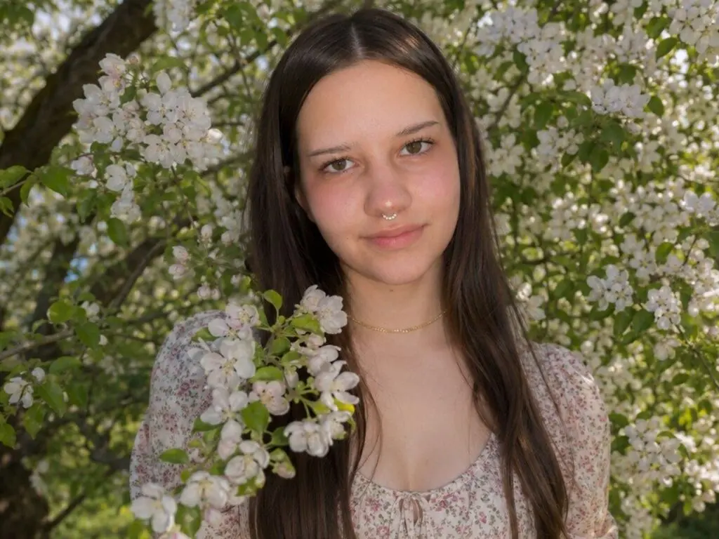 Young woman in a floral print dress smiling amidst blooming white apple blossoms in a sunny garden.