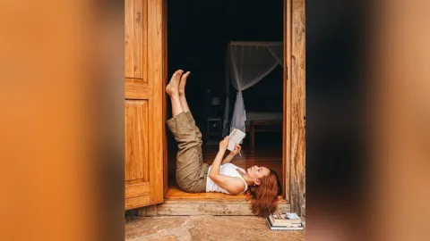 MilaSwan — MilaSwan lies on a wooden floor in a white tank top and tan pants, holding a book with her legs raised in the air.