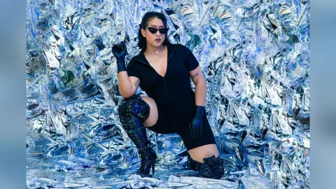 KimRoberth poses in a black bodysuit and black gloves against a shiny silver tinfoil background, wearing dark sunglasses.