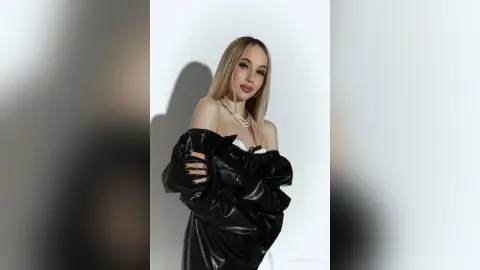 MarryDavies poses in a black leather off-the-shoulder top, looking at the camera against a white background.
