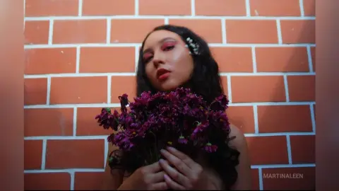 Young woman with dark hair holding a bouquet of purple flowers against a red brick wall in soft lighting.