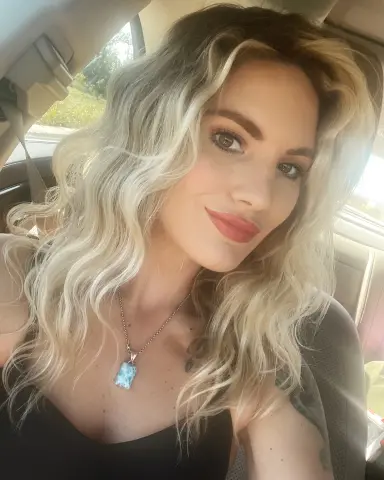 GoddessBiancaBelle smiles for a selfie in her car, showing off her long blonde wavy hair and a blue pendant necklace.