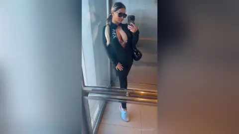 Stylish woman in a black zip-up jacket and leggings taking a confident mirror selfie in a modern hallway.