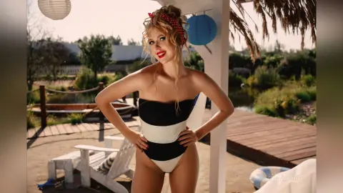 Blonde woman in a black and white swimsuit posing on a sunny wooden deck by a tropical garden.
