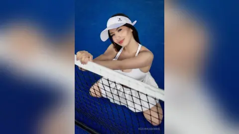 Smiling woman in white tennis outfit and visor leaning on a net against a blue background.