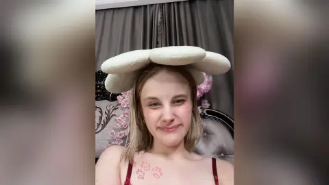 Smiling young woman wearing a large white puffy cloud headband in a cozy room with pink floral decor.