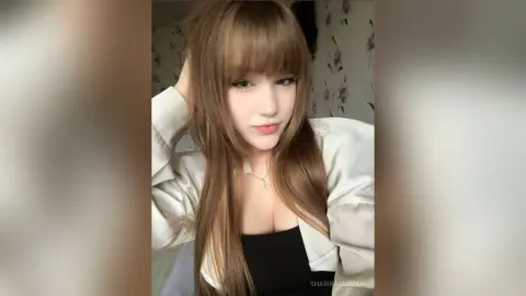 SharikaGlidden poses with long brown hair and bangs, wearing a white jacket over a black top and a silver cross necklace.
