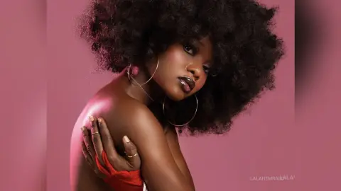 Beautiful Black woman with voluminous afro hair and gold hoop earrings posing against a pink background.