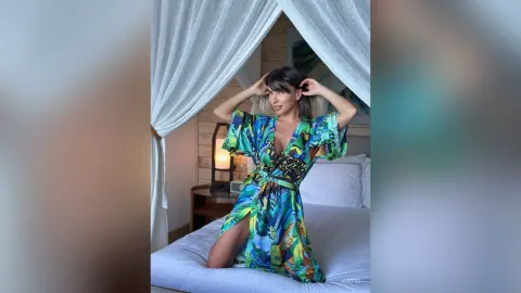 Katia poses on a bed wearing a colorful patterned wrap dress with her hands in her hair.