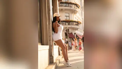 NicoleBella leans against a stone wall wearing a white sleeveless bodysuit and a white baseball cap.
