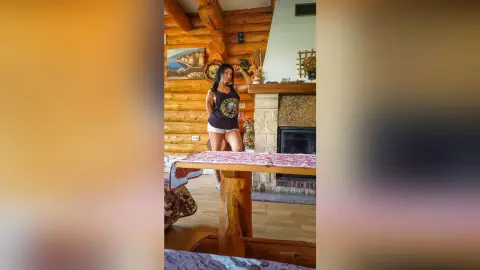 MissyJolie stands in a wood-paneled room wearing a black tank top and white shorts.