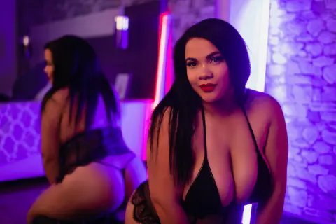 Grace_Clarck poses in a black lace bra and panties, showing her cleavage and bare back against a purple-lit stone wall.