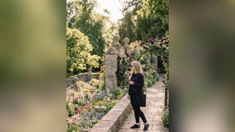 Blonde woman in a black outfit standing in a lush summer flower garden by a stone archway.