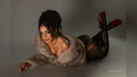 Curvy woman in a cozy faux fur coat and patterned tights lounging on a grey studio floor with a glamorous mood.