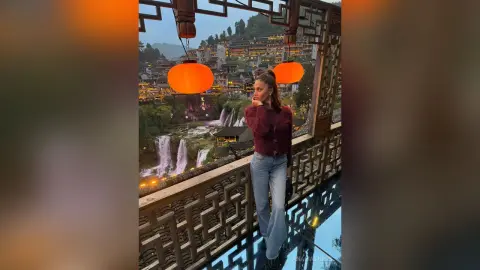 Thoughtful woman in a maroon sweater on a traditional balcony overlooking a scenic waterfall at dusk.