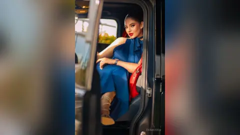 Elegant woman in a blue sleeveless dress sitting in a vintage car doorway during golden hour.