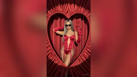 Kalira poses in a red heart-shaped curtain opening, wearing a red cut-out bodysuit and long red gloves with sunglasses.