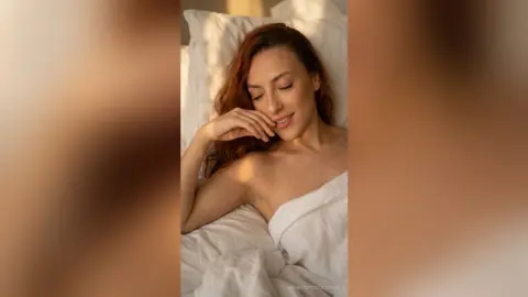 Smiling woman relaxing in white bed linens under warm sunlight for a peaceful morning mood.