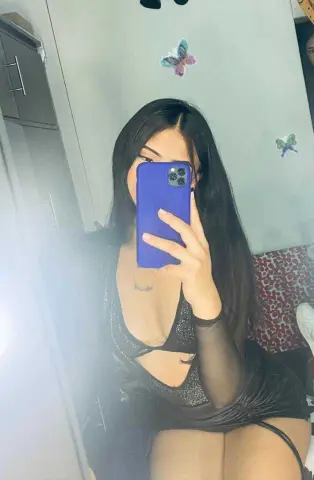 Young woman in a black sparkly cutout dress taking a mirror selfie in a bedroom setting.
