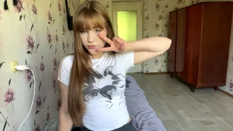 SharikaGlidden poses with a peace sign, wearing a white graphic t-shirt and showing her long brown hair with bangs in a room with floral wallpaper.