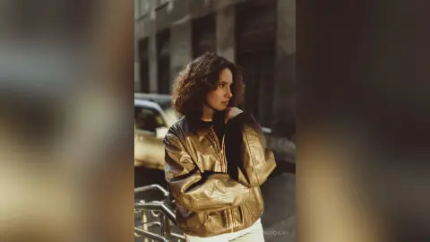 Young woman in a metallic gold bomber jacket standing on a city street during golden hour.