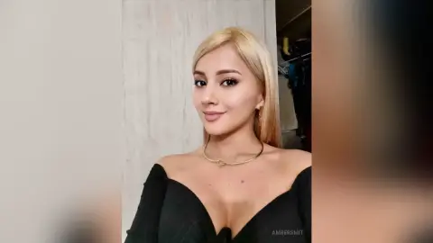 Blonde woman in a black off-the-shoulder top and gold necklace posing with a soft smile indoors.