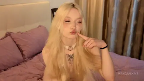 Blonde woman with long hair and heart choker posing playfully on a purple bed in a cozy bedroom setting.