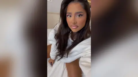 DayanaNoir — Young woman in a white cozy bathrobe relaxing in a bright bedroom with a serene mood.
