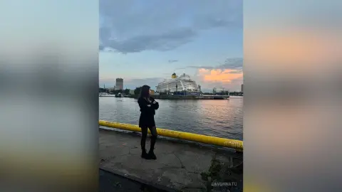 Woman in black outfit standing by a river at sunset with a large cruise ship in the background.